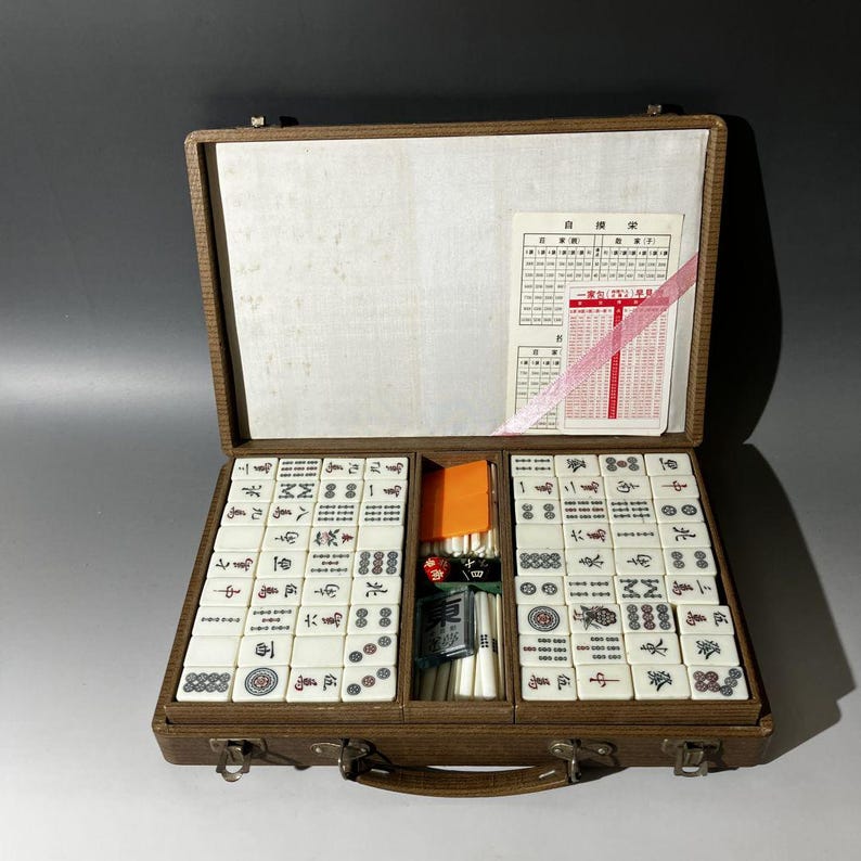 Puede incluir: Un juego de mahjong vintage en un malet&iacute;n marr&oacute;n texturizado con cierres met&aacute;licos. El set incluye fichas blancas con caracteres grabados en negro, rojo y verde, un compartimento central con varillas de conteo blancas, un dado rojo y una tarjeta.