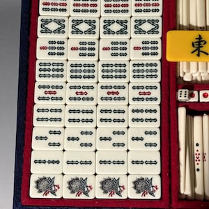 May include: A vintage Mahjong set with ivory-colored tiles, featuring black and red symbols and characters. The set includes dice and a yellow tile with a black character. The tiles are arranged in a blue and red velvet-lined case.