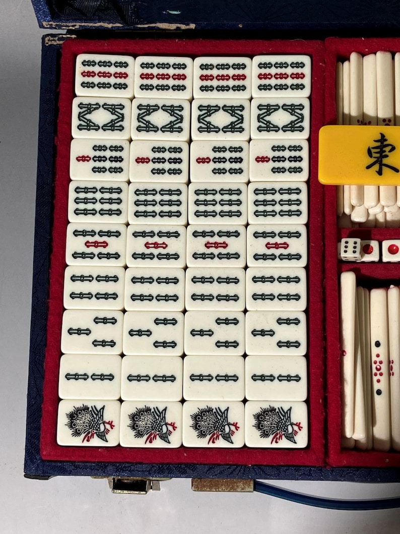 May include: A vintage Mahjong set with ivory-colored tiles, featuring black and red symbols and characters. The set includes dice and a yellow tile with a black character. The tiles are arranged in a blue and red velvet-lined case.
