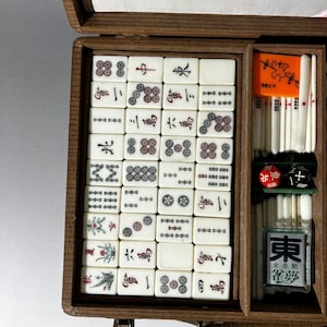 Puede incluir: Una caja de madera que contiene un juego de fichas de Mahjong de color blanco roto con caracteres y s&iacute;mbolos grabados en negro, rojo y verde. El juego incluye una ficha naranja brillante con una ilustraci&oacute;n de un p&aacute;jaro, varas blancas y dos dados.