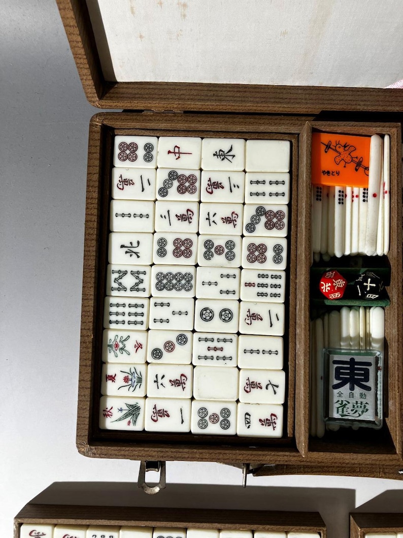 Puede incluir: Una caja de madera que contiene un juego de fichas de Mahjong de color blanco roto con caracteres y s&iacute;mbolos grabados en negro, rojo y verde. El juego incluye una ficha naranja brillante con una ilustraci&oacute;n de un p&aacute;jaro, varas blancas y dos dados.