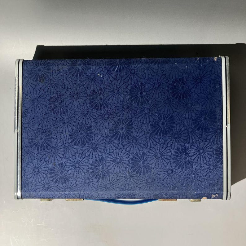 May include: A rectangular, navy blue box with a floral pattern. The box has a silver trim and a blue handle. The floral pattern is a repeating design of small, stylized flowers. The box appears to be closed.