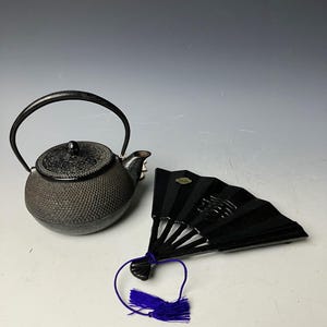 以下が含まれることがあります： 黒色の鋳鉄製の急須で、表面はざらざらしています。黒色の鋳鉄製の扇形の鍋敷きで、紫色の房が付いています。