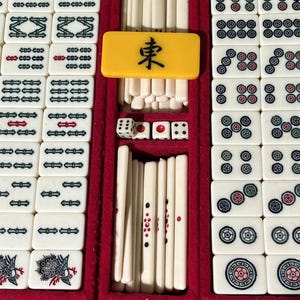 May include: A vintage Mahjong set in a blue case with a red velvet interior. The set includes white tiles with black, red, and green markings, dice, and a yellow tile with a black character. The tiles are arranged in rows.