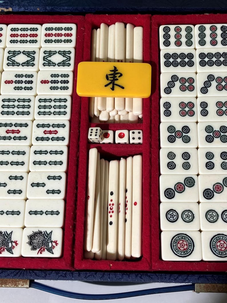 May include: A vintage Mahjong set in a blue case with a red velvet interior. The set includes white tiles with black, red, and green markings, dice, and a yellow tile with a black character. The tiles are arranged in rows.