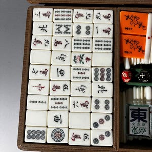 Puede incluir: Una caja de madera que contiene un juego de fichas de Mahjong de color blanco hueso con caracteres, s&iacute;mbolos y dise&ntilde;os florales en tinta negra, roja y verde. El juego incluye marcadores rectangulares naranjas con ilustraciones de aves.