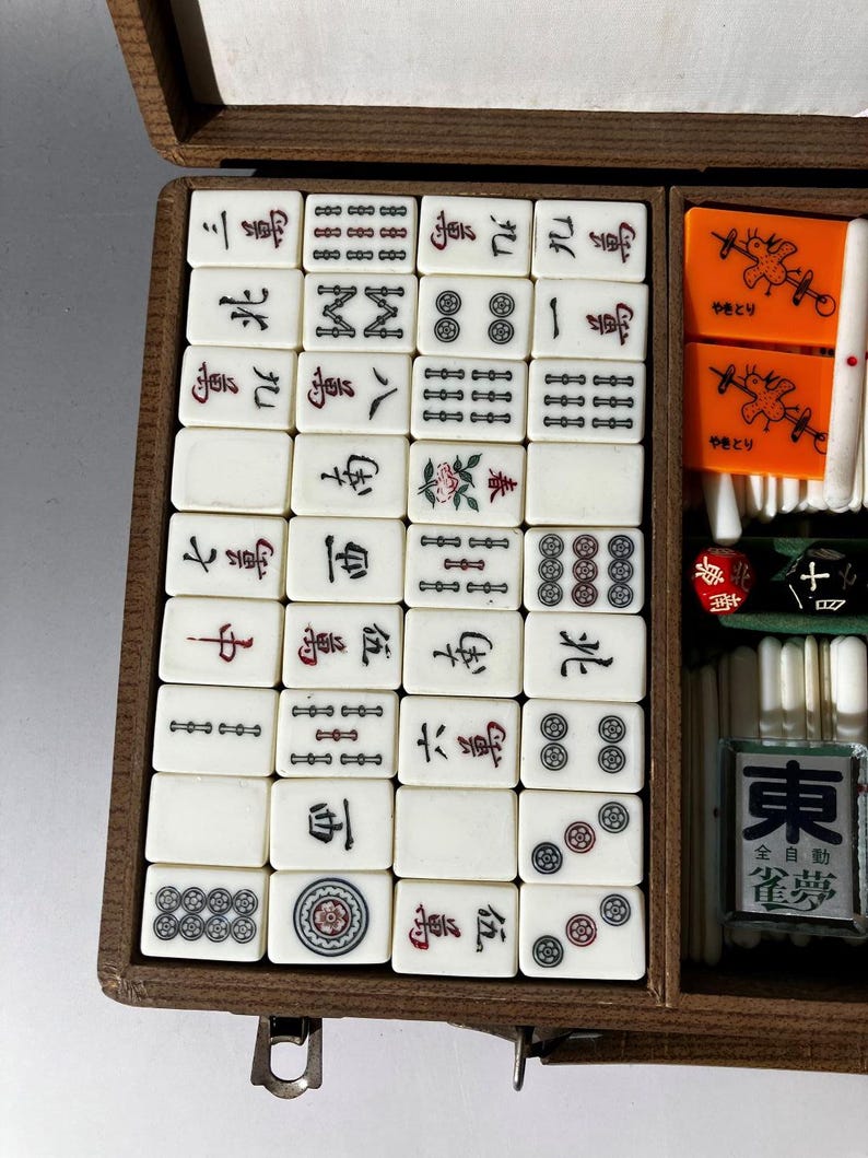 Puede incluir: Una caja de madera que contiene un juego de fichas de Mahjong de color blanco hueso con caracteres, s&iacute;mbolos y dise&ntilde;os florales en tinta negra, roja y verde. El juego incluye marcadores rectangulares naranjas con ilustraciones de aves.