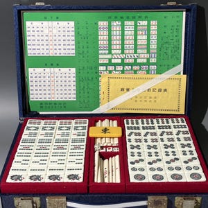 May include: Open blue case containing a Mahjong set. The set includes ivory-colored tiles with red and green markings, dice, and a yellow tile marked with the character for "East". The case also holds charts and a booklet.