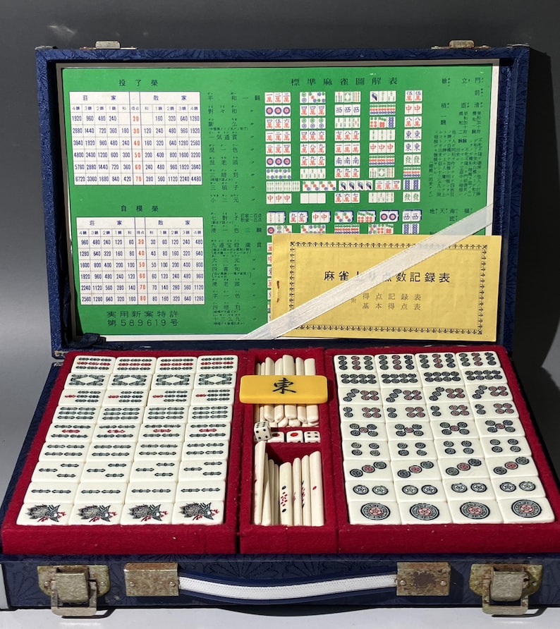 May include: Open blue case containing a Mahjong set. The set includes ivory-colored tiles with red and green markings, dice, and a yellow tile marked with the character for "East". The case also holds charts and a booklet.