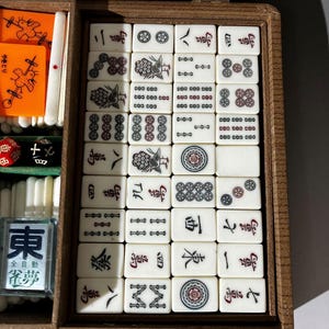 Puede incluir: Una caja de madera que contiene un juego de fichas de Mahjong con varios s&iacute;mbolos, caracteres y patrones grabados en negro y rojo. El conjunto incluye fichas rectangulares blancas, fichas naranjas con ilustraciones de aves y un dado negro.