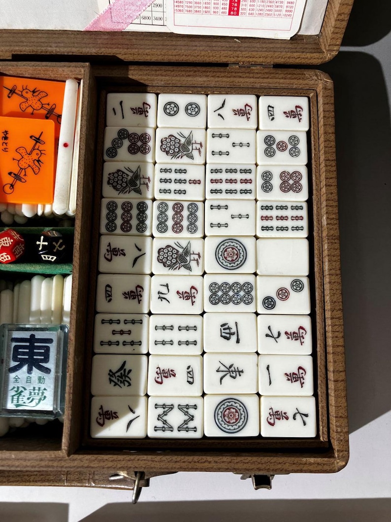 Puede incluir: Una caja de madera que contiene un juego de fichas de Mahjong con varios s&iacute;mbolos, caracteres y patrones grabados en negro y rojo. El conjunto incluye fichas rectangulares blancas, fichas naranjas con ilustraciones de aves y un dado negro.