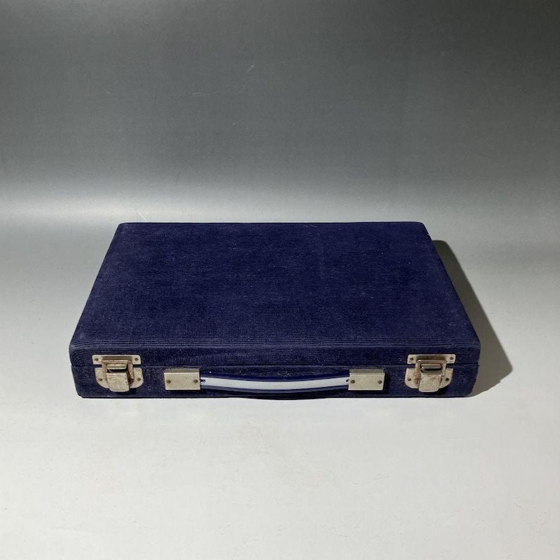 Pu&ograve; includere: Una valigetta rettangolare rivestita in velluto blu scuro, dotata di una maniglia centrale con un dettaglio a righe bianche e blu. La custodia &egrave; fissata da due chiusure in metallo color argento sul bordo anteriore, con un aspetto piatto.