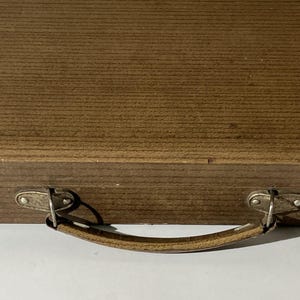 Puede incluir: Un estuche de madera marr&oacute;n con una textura de veta horizontal y un asa curva sujeta con herrajes de metal plateado. El asa est&aacute; asegurada por dos placas de metal, cada una fijada con dos peque&ntilde;os remaches al frente de la caja.