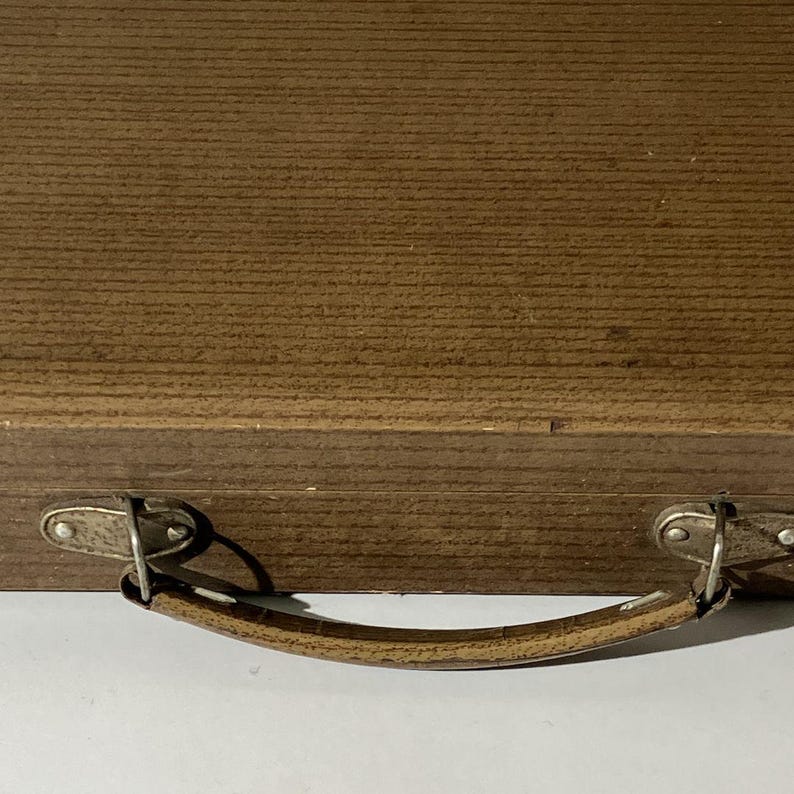 Puede incluir: Un estuche de madera marr&oacute;n con una textura de veta horizontal y un asa curva sujeta con herrajes de metal plateado. El asa est&aacute; asegurada por dos placas de metal, cada una fijada con dos peque&ntilde;os remaches al frente de la caja.
