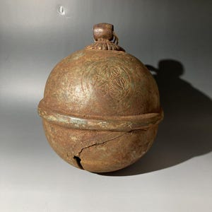 May include: A weathered, spherical metal bell with a loop at the top and a decorative floral pattern. The bell has a crack running down the side, and a rusty, aged appearance. The bell is likely an antique.