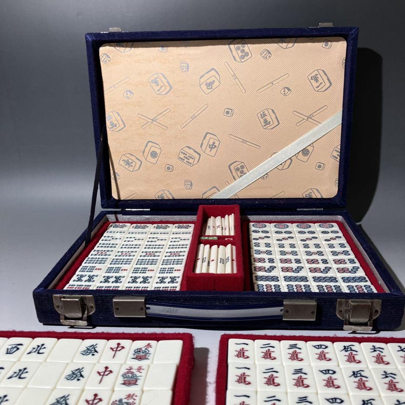 Pu&ograve; includere: Una custodia per mahjong rivestita in velluto blu contenente tessere bianche con caratteri rossi, blu e neri. Il coperchio interno presenta un motivo beige di tessere, e uno scomparto centrale rosso contiene bastoncini segnapunti bianchi.