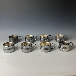 以下が含まれることがあります： 小さな取っ手が付いた、銀色のカップが8つ。カップの外側はハンマーで叩いたような質感で、内側は光沢のある銀色です。白い面に並べられています。