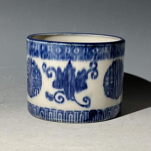 Può includere: Un contenitore cilindrico in ceramica con base bianca e motivi decorativi blu. Il design include motivi floreali e bordi geometrici. Il contenitore ha un diametro di circa 10 cm e un'altezza di 7,6 cm.