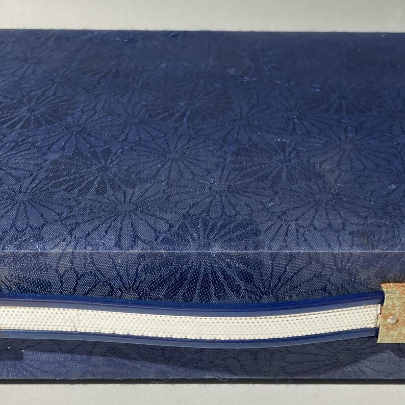 May include: A close-up of a navy blue box with a floral pattern. The box is closed, with a white and blue trim along the edge. A small, rusty metal clasp is visible on the side.