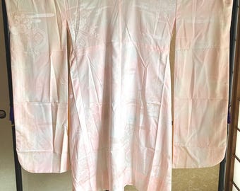 Könnte beinhalten: Ein hellrosa Kimono mit einem dezenten Blumen- und geometrischen Muster. Das traditionelle japanische Kleidungsstück wird auf einem schwarzen Rahmen präsentiert, der seine langen Ärmel und sein fließendes Design zeigt. Der Stoff wirkt weich und leicht.