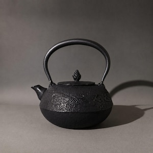 Puede incluir: Una tetera de hierro fundido negro con una superficie texturizada y diseños de peces koi en relieve en el cuerpo. La tetera tiene un asa curva, un pico pequeño y una tapa extraíble rematada con un pequeño pomo decorativo en el centro.