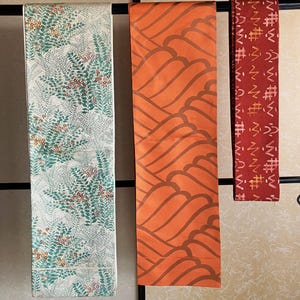 Può includere: Tre pannelli di tessuto decorativi appesi a un'asta nera. I pannelli presentano diversi motivi: un pannello bianco con foglie verdi e bacche rosse, un pannello arancione con forme ondulate marroni e un pannello rosso con forme geometriche bianche.