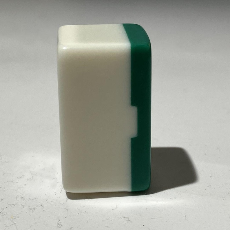 Pu&ograve; includere: Una tessera rettangolare da mahjong posizionata in verticale, caratterizzata da una faccia anteriore bianca opaca e liscia e da un retro di colore verde scuro uniforme. La tessera presenta angoli arrotondati, finitura lucida e una giuntura.
