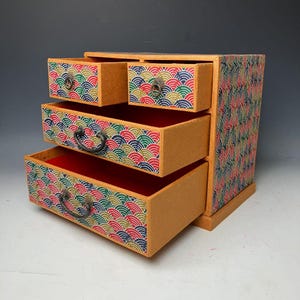Puede incluir: Un pequeño mueble de cajones de madera con tres cajones extraíbles. Los cajones y los lados están decorados con un colorido patrón de abanicos repetido en rojo, azul, verde y dorado. Los interiores de los cajones son rojos y los tiradores son de metal oscuro.