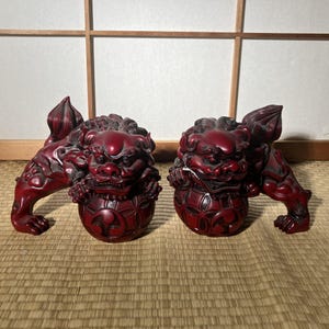 以下が含まれることがあります： 2つの赤いFoo Dogの像。各像はライオンのような頭、詳細な鬣、装飾的なボールを持っています。像はしゃがんだ姿勢で、前足はボールの上にあります。背景は明るい色のスクリーンです。