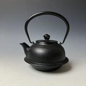 Könnte beinhalten: Eine schwarze gusseiserne Teekanne mit gebogenem Griff und kleinem Ausguss. Die Teekanne hat eine strukturierte Oberfläche und einen dekorativen Knopf auf dem Deckel. Die Teekanne steht auf einem runden Sockel. Ein traditioneller Küchenartikel.