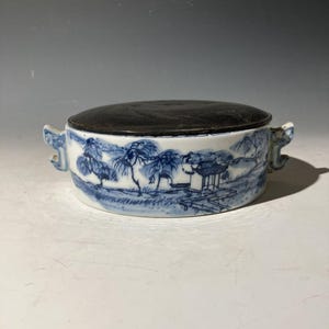 以下が含まれることがあります： 暗い蓋が付いた、白と青の丸い陶器の容器。容器には、木や建物のある風景のデザインが施されています。両側に2つの装飾的なハンドルが付いています。