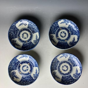 以下が含まれることがあります： 青い花柄の小さな白い陶器の皿が4枚セットになっています。皿は白い背景に青い花柄が描かれており、中央には金色の模様があります。