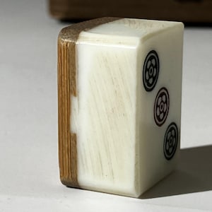 Puede incluir: Una ficha de mahjong rectangular de dos tonos, con una cara lisa de color blanco hueso y un reverso de bamb&uacute; texturizado en color marr&oacute;n claro. La cara blanca muestra tres s&iacute;mbolos florales circulares dispuestos verticalmente en tinta.