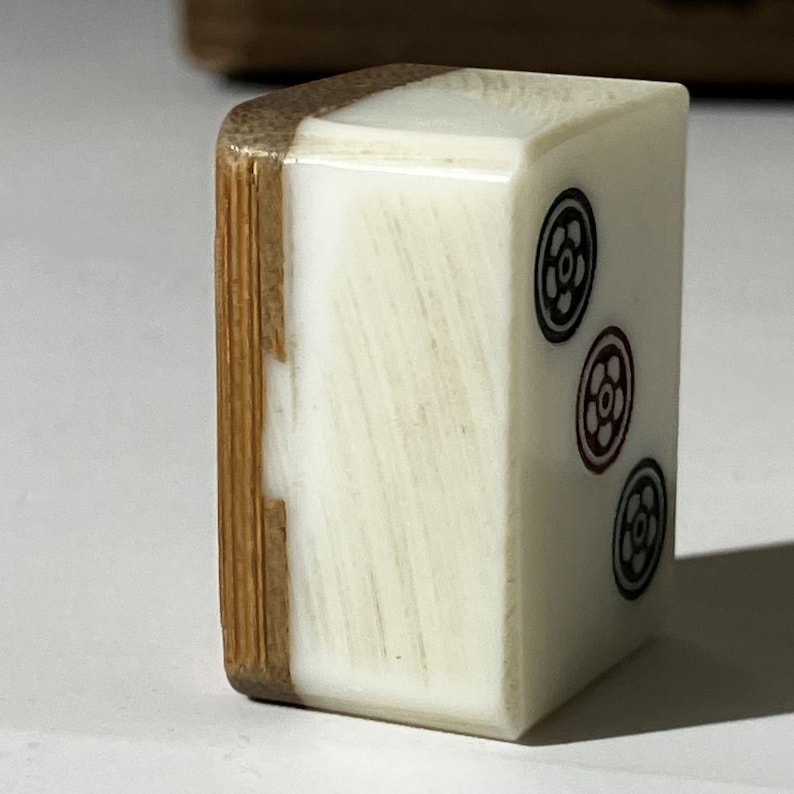 Puede incluir: Una ficha de mahjong rectangular de dos tonos, con una cara lisa de color blanco hueso y un reverso de bamb&uacute; texturizado en color marr&oacute;n claro. La cara blanca muestra tres s&iacute;mbolos florales circulares dispuestos verticalmente en tinta.