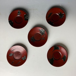 以下が含まれることがあります： 黒い花柄の赤い漆塗りの日本製茶碗5個セット。茶碗は丸く、中央に茶碗を置くための小さなへこみがあります。