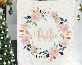May include: A white fleece blanket with a pink polka dot pattern. A floral wreath with pink and orange flowers surrounds the letter "M" and the name "Maddie" in gold lettering.