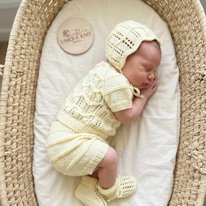 Puede incluir: Un bebé dormido con un conjunto de punto amarillo pálido, que incluye un gorro, una camiseta de manga corta, pantalones cortos y botines. El bebé está acunado en una cesta tejida. Disco de madera con el texto "UNIQUE KNIT Designs".