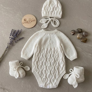 May include: A cream-coloured knitted long-sleeve romper featuring a vertical leaf-patterned texture, accompanied by a matching bonnet with tie straps and two booties. A circular wooden tag displays the text UNIQUE KNIT DESIGNS.
