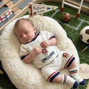 May include: A sleeping baby wearing a cream-colored knitted outfit with red and blue accents. The outfit includes a top with the word "England" and a floral emblem, shorts, and socks. The baby is lying on a fluffy white surface, with a miniature soccer field in the background.