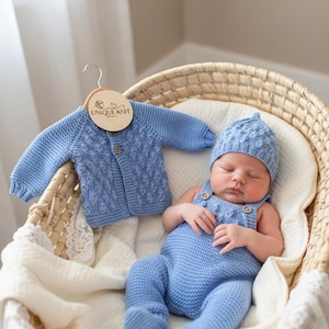 May include: A baby wearing a light blue knitted outfit, including a hat and overalls, lies in a wicker basket. A matching blue cardigan hangs nearby. The cardigan has a wooden tag that says "UNIQUE KNIT".