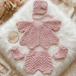 Op de afbeelding: Een babykleding set in zachtroze, bestaande uit een vest, shorts, muts en babyschoentjes. Het breiwerk heeft een chevronpatroon en knoopsluitingen. De set wordt gepresenteerd op een witte, pluizige ondergrond.