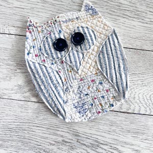 May include: A quilted fabric owl ornament featuring a patchwork design of blue and white stripes mixed with small floral patterns. The owl has two dark navy blue circular buttons for eyes and a stitched triangular beak, with a textured quilted finish.
