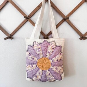 K&ouml;nnte beinhalten: Eine wei&szlig;e Canvas-Tragetasche mit einem lila und gelben Blumendesign. Die Blume besteht aus Patchwork-Stoff und hat eine gelbe Mitte.