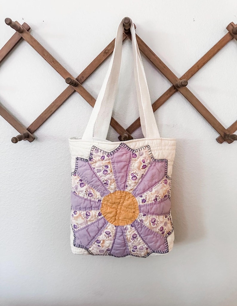 K&ouml;nnte beinhalten: Eine wei&szlig;e Canvas-Tragetasche mit einem lila und gelben Blumendesign. Die Blume besteht aus Patchwork-Stoff und hat eine gelbe Mitte.