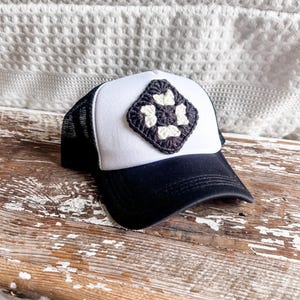 May include: A black and white crocheted square patch is sewn onto a white trucker hat with a black mesh back. The hat is sitting on a weathered wooden surface.