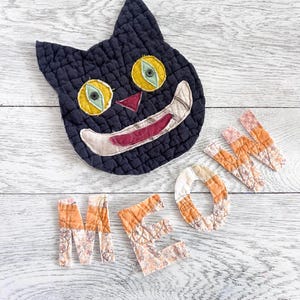 May include: A quilted black cat face with yellow eyes, green pupils, and a wide grin, accompanied by fabric letters spelling MEOW. The letters are crafted from orange and floral patterned quilted fabric, arranged in a playful, decorative layout.