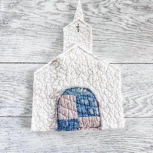 May include: A white quilted fabric ornament shaped like a church with a steeple and a small black embroidered cross. The center features an arched doorway constructed from a patchwork of blue and light pink textured fabric squares.