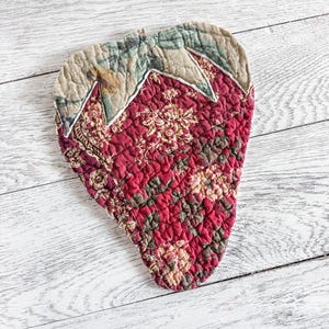 May include: A strawberry-shaped quilted fabric pad featuring a deep red base with a dark floral pattern and a beige top section with a green leaf print. The item has visible stitching throughout the quilted surface and a pointed bottom edge.