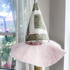 May include: A quilted witch hat featuring a tall, conical crown made of sage green fabric with a floral pattern and a white square patch. The wide, circular brim is solid light pink with a swirling quilted texture and a white finished edge.
