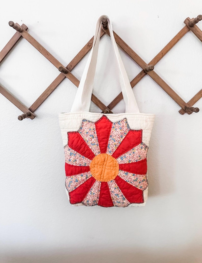 K&ouml;nnte beinhalten: Eine wei&szlig;e Canvas-Tragetasche mit einem roten, orangefarbenen und blauen Blumendesign. Die Tasche hat zwei lange Henkel und h&auml;ngt an einem Holzgestell.